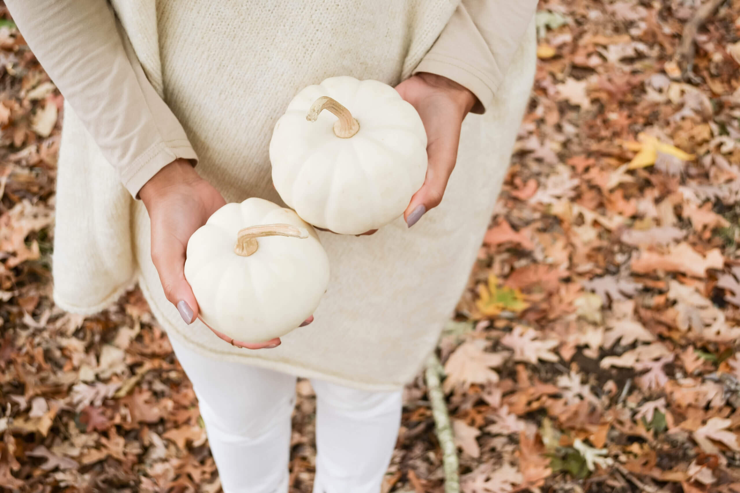 woman in white pants and cream top holding 2 white pumpkins